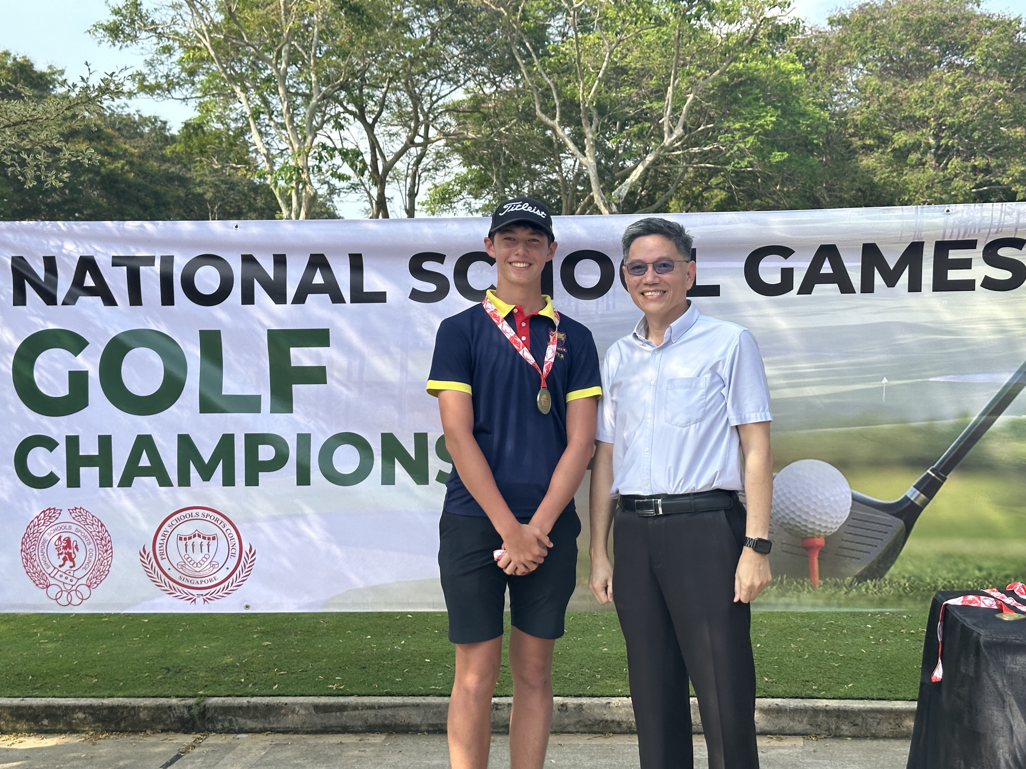 A young golfer wearing a medal poses with an older man in front of a National School Games Golf Championship banner.